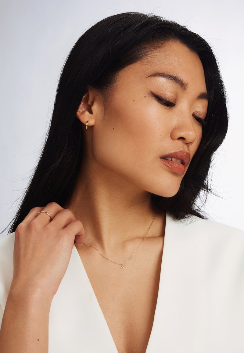 Boucles d'oreilles en or, collier délicat en or avec un pendentif « A », peau lisse et cheveux noirs, portant un haut blanc à col en V.