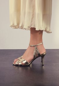 Sandali con tacco alto color oro metallico con un design a cinturini, che mostrano una punta aperta e una cavigliera, contro una gonna plissettata color crema.