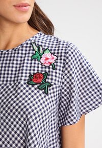 Blå och vit rutig topp med två blommönstrade lappar i rosa och rött med gröna blad, korta ärmar och rund halsringning.