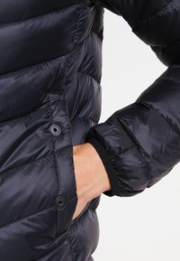 Svart puffjacka med kviltad design, utrustad med en sidoficka med tryckknappslås och elastiska ärmslut för en säker passform.