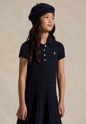 Fille portant une robe en maille torsadée bleu marine avec des boutons et un béret assorti, debout devant un fond beige uni.