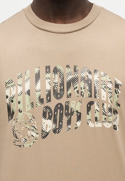 Άνω μέρος σώματος που φοράει μπεζ πουκάμισο με μεγάλο κείμενο σε σχέδιο καμουφλάζ "BILLIONAIRE BOYS CLUB" και γραφικό με κράνος αστροναύτη στο μπροστινό μέρος.