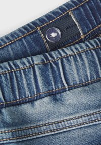 Pantalons en denim de couleur bleu décoloré, avec des détails en couture et un bouton visible. La taille est dotée de fronces élastiques pour un confort optimal.