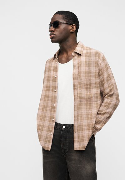 Άνδρας με μαύρα γυαλιά ηλίου, μπεζ καρό ανοιχτό πουκάμισο πάνω από λευκό φανελάκι και σκούρο τζιν, στέκεται με το ένα χέρι στην τσέπη.