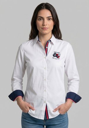 Mujer que lleva una camisa blanca abotonada con ribetes a rayas en azul marino y rojo, escudo bordado en el pecho y puños azul marino, de pie con las manos en los bolsillos.