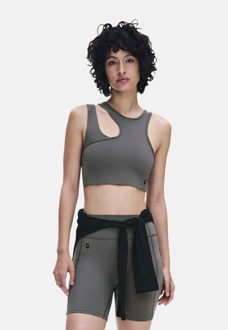Jeune femme aux cheveux bouclés porte un crop top asymétrique gris et un short de cycliste taille haute assorti avec un sweat-shirt noir noué autour de la taille.