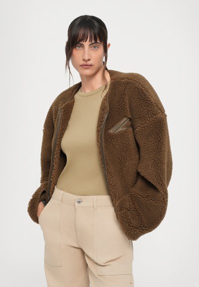 Braune Fleecejacke mit oversized Design, ausgestattet mit einem Reißverschluss und Taschen. Getragen über einem beigen, enganliegenden Shirt und hellbeigen Hosen.