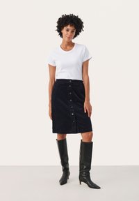 Fekete kordbársony gombos szoknya fehér pólóval és térd fölé érő fekete csizmával párosítva, kiemelve a testhezálló siluettet és a texturált anyagot.