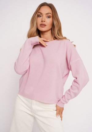 Kobieta o długich blond włosach, ubrana w jasnoróżowy sweter i kremowe spodnie, pozująca z jedną ręką przy szyi, a drugą na biodrze.