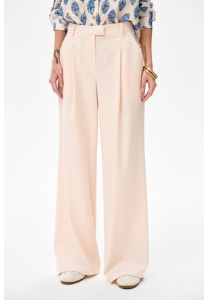 Kobieta ubrana w szerokie, jasnoróżowe spodnie z zakładkami z przodu, zestawione z białymi butami i niebieską kwiecistą koszulą wpuszczoną do środka.