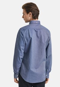 Heren overhemd met lange mouwen van blauwe chambray stof. Heeft een button-down kraag, een gebogen zoom en knoopmanschetten. Vloeiende textuur met een subtiele glans.