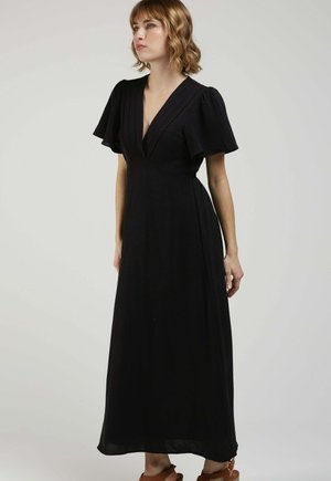 Femme debout portant une longue robe noire à manches évasées et un décolleté en V profond, associée à des sandales compensées marron.