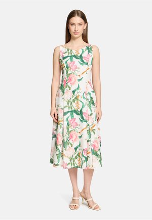 Frau in ärmellosem, weißem Midikleid mit pink-grünem Blumendruck, steht aufrecht mit beigen Sandalen mit Absatz vor einfarbigem Hintergrund.