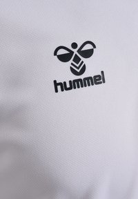 Vit texturerad tyg med en svart Hummel-logotypdesign som har stiliserade bi-element och ett unikt geometriskt mönster i mitten.