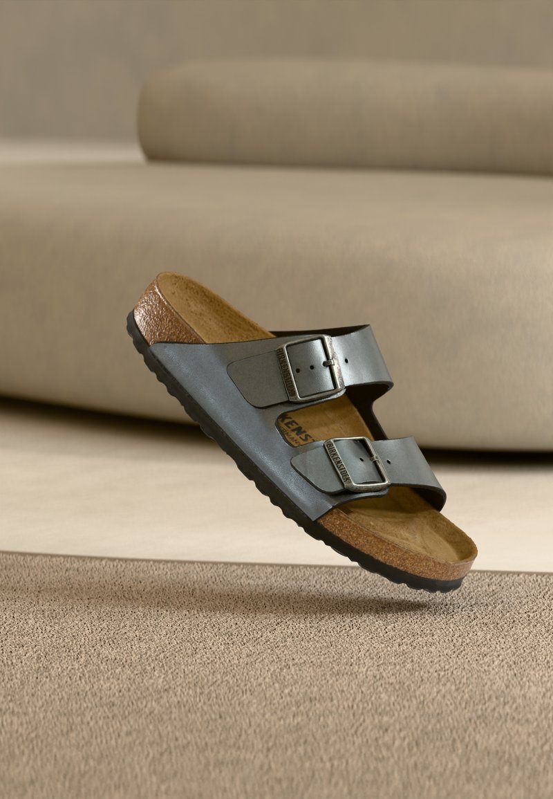 Μαύρο σανδάλι με δύο λουριά και ρυθμιζόμενες μεταλλικές αγκράφες, πάτο από φελλό και υφασμένο πέλμα, το οποίο βρίσκεται πάνω σε μπεζ χαλί σε δωμάτιο με ουδέτερους τόνους.