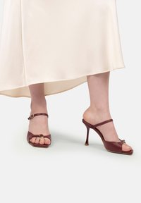 Picioare feminine purtând sandale cu toc înalt burgund cu bretele la glezne, stând sub o fustă midi de culoare crem pe un fundal alb.