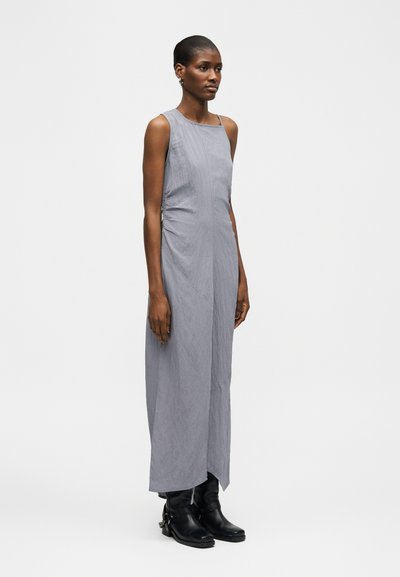 Ψηλή γυναίκα που φοράει αμάνικο γκρι φόρεμα μέχρι τον αστράγαλο και μαύρα μποτάκια, στέκεται μπροστά σε λευκό, απλό φόντο.