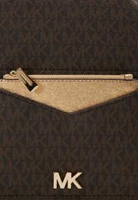 Sac de créateur marron foncé texturé avec un motif MK répété, poche avant zippée dorée et logo MK doré en bas au centre.