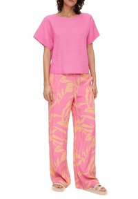 Donna che indossa una maglietta rosa morbida a maniche corte con pantaloni larghi rosa caratterizzati da motivi di foglie tropicali beige e sandali beige.