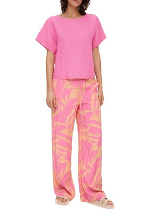 Donna che indossa una maglietta rosa morbida a maniche corte con pantaloni larghi rosa caratterizzati da motivi di foglie tropicali beige e sandali beige.