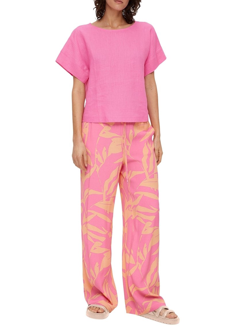 Donna che indossa una maglietta rosa morbida a maniche corte con pantaloni larghi rosa caratterizzati da motivi di foglie tropicali beige e sandali beige.