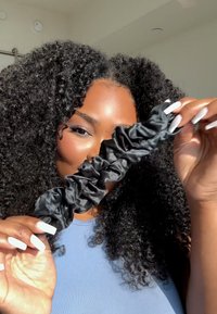 Scrunchie nera in satin tenuta tra le mani, caratterizzata da una trama arricciata e una superficie liscia. Lunghe e morbide onde incorniciano lo sfondo.
