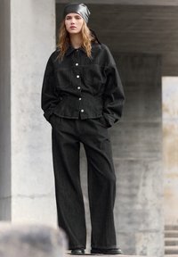 Czarna kurtka dżinsowa z przednimi kieszeniami, zapinana na srebrne guziki, w połączeniu z pasującymi szerokimi spodniami. Modelka ma na sobie czarną satynową chustę na głowie.