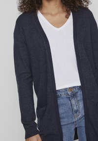 Marinblå stickad cardigan med öppen front, långa ärmar och två framfickor, över en vit V-ringad t-shirt och blå jeans.