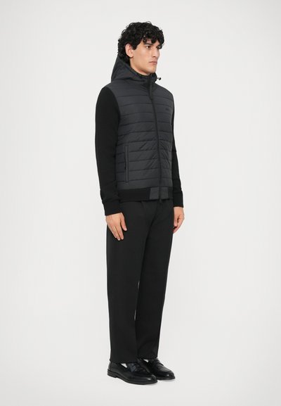 Colete acolchoado preto com capuz, apresentando painéis acolchoados, fecho de correr e bolsos frontais; usado sobre uma camisola preta de manga longa e calças escuras.