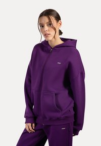 Sudadera con cremallera en color púrpura, con una gran capucha, bolsillos tipo canguro y puños acanalados. Fabricada con material suave, presenta un pequeño logo de la marca en el pecho.