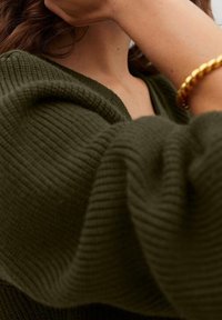 Pull en maille côtelée vert olive avec un col en V profond. Bracelet doré texturé visible au poignet. Le tissu semble doux et légèrement drapé.