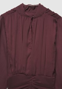 Blusa color marrone con colletto alto, chiusura con bottoni, davanti plissettato e arricciature in vita. Realizzata in tessuto leggero e testurizzato.