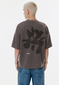 Mand med kort, lys blondt hår iført en oversized mørkegrå t-shirt med abstrakt sort print og tekst, parret med blå jeans, vender bort.