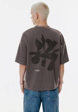 Moški s kratkimi blond lasmi nosi prevelik temnosiv T-shirt z abstraktnim črnim vzorcem in besedilom, kombiniran z modrimi kavbojkami, obrnjeno stran.