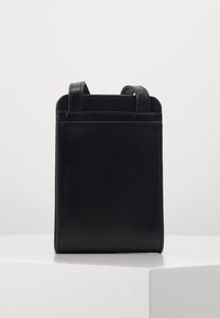 Malá crossbody taška z černé hladké kůže s dvěma úzkými popruhy, stojící vzpřímeně na bílé ploše.