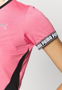 Rosa sporttröja med en texturerad yta. Svarta detaljer på sidorna och ett svart band med "PUMA"-text på ärmen. Silverfärgad logotyp på framsidan.