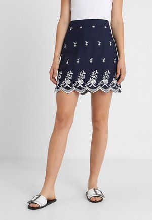 Kobieta ubrana w granatową mini spódnicę z białym kwiatowym haftem i falowanym brzegiem, zestawioną z białymi klapkami.