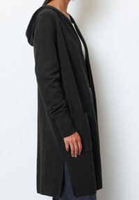 Lång svart hoodie cardigan, tillverkad av mjukt tyg, med sidofickor och sprund i fållen för ökad rörlighet.