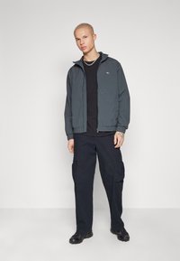 Chaqueta bomber con capucha en gris oscuro, con puños y cintura elásticos, llevada sobre una camiseta negra y combinada con pantalones cargo negros.