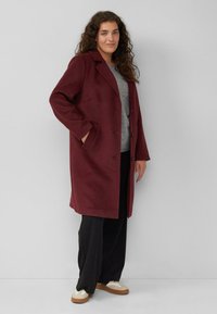 Vrouw die staat met haar hand in de zak, gekleed in een lange bordeauxrode jas, grijze top, zwarte wijde broek en beige sneakers tegen een grijze achtergrond.