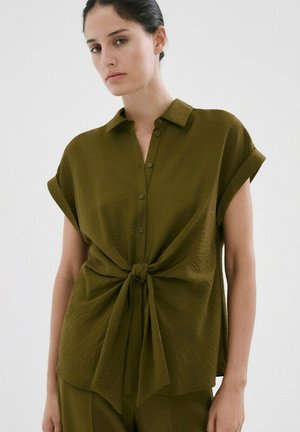 Femme portant une chemise à manches courtes boutonnée couleur vert olive nouée à la taille, debout devant un fond blanc uni.