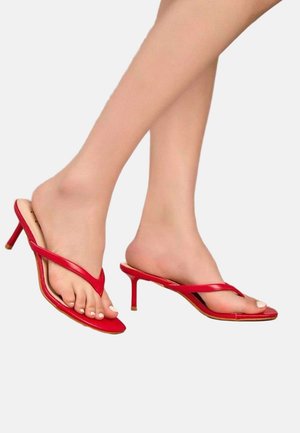 Pieds portant des sandales rouges à talons hauts avec de fines brides sur un fond clair uni.