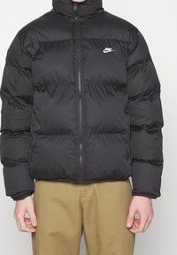 Chaqueta de plumas negra de Nike con cremallera frontal y logotipo en el pecho, combinada con pantalones marrón claro, foto que muestra el torso y los brazos.