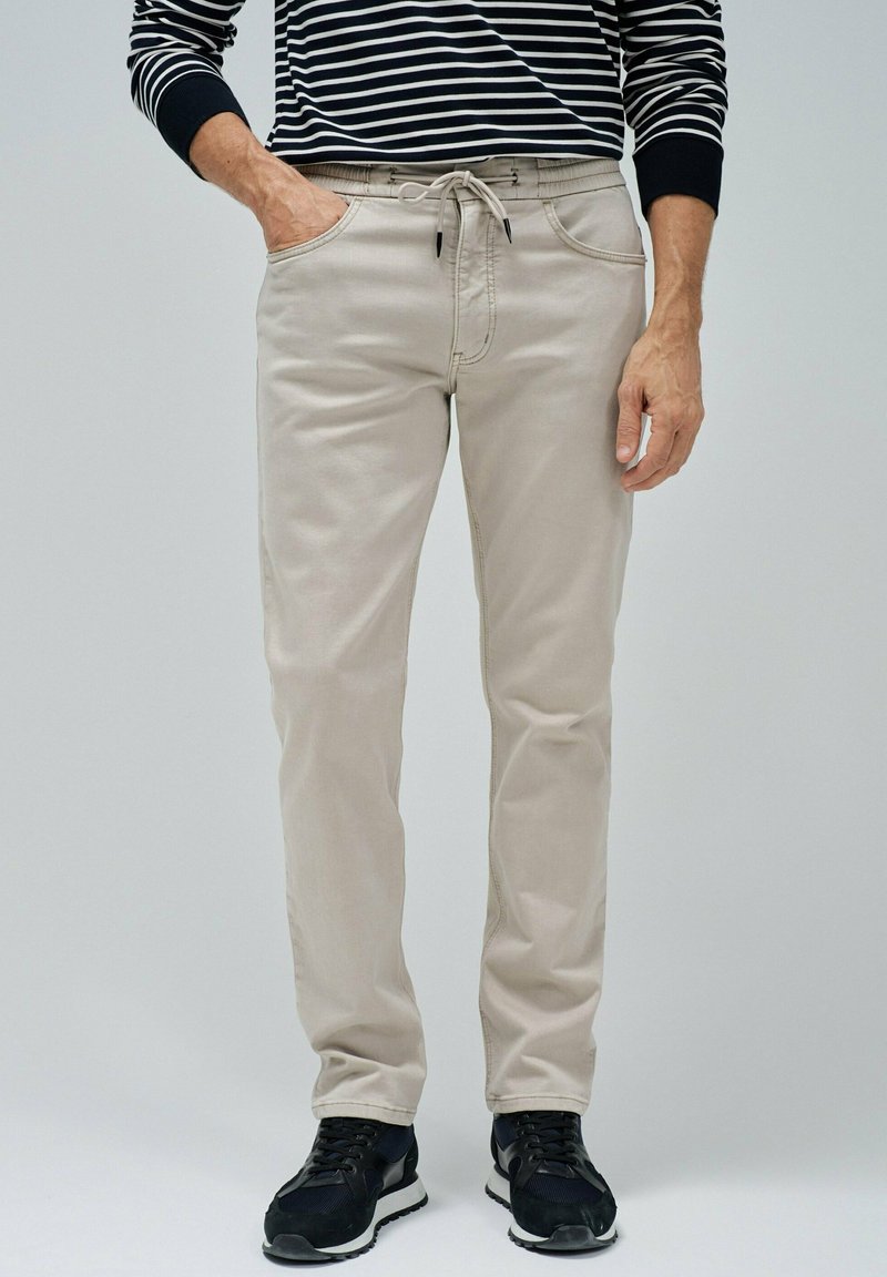 Pantalones de mezcla de algodón beige con cintura ajustable, dos bolsillos laterales y corte slim, combinados con zapatillas deportivas negras.