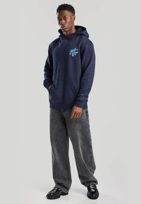 Sweatshirt bleu marine avec poche kangourou, poignets côtelés et impression sur le devant. Porté avec un jean large gris et des chaussures noires à semelles épaisses.