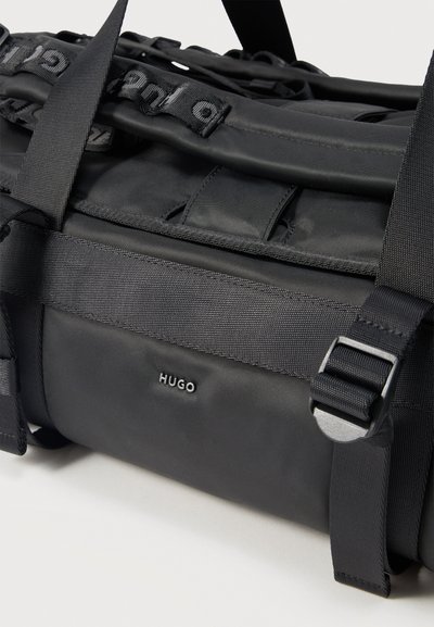 Черен сак Hugo тип duffel с регулируема презрамка и брандиран текстилен детайл на бял фон.