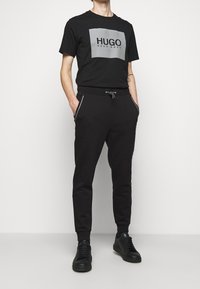 Schwarzes Baumwoll-T-Shirt mit grauem rechteckigem Logo. Kombiniert mit schwarzen Jogginghosen, die Seitentaschen und einen elastischen Bund haben. Schwarze Sneakers.