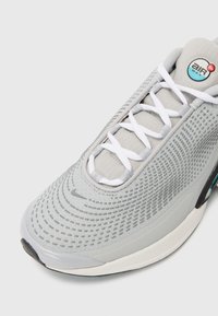 Harmaa lenkkari, jossa on teksturoitu verkko-yläosa, valkoiset nauhat ja näkyvä Air Max -vaimennusyksikkö. Kengässä on pyöreä logo kielen kohdalla.