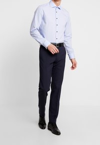 Vaaleansininen nappikauluspaita, jossa on levitetty kaulus, mustat napit ja pitkät hihat, yhdistettynä tummansinisiin housuihin ja mustiin juhlakengiin.