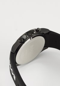 Black rubber strap watch featuring a circular case, two push buttons, textured accents, and a smooth silver caseback with branding.